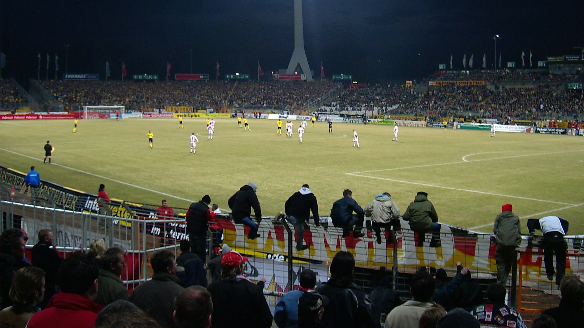 Fans des 1. FC Köln sitzen auf dem Zaun zum Innenraum des Rudolf-Harbig-Stadions und verfolgen das Spiel bei Dynamo Dresden.