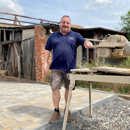 Ein Mann steht in einem Hof an einer Säge, die durch vieles Arbeiten eingestaubt ist.