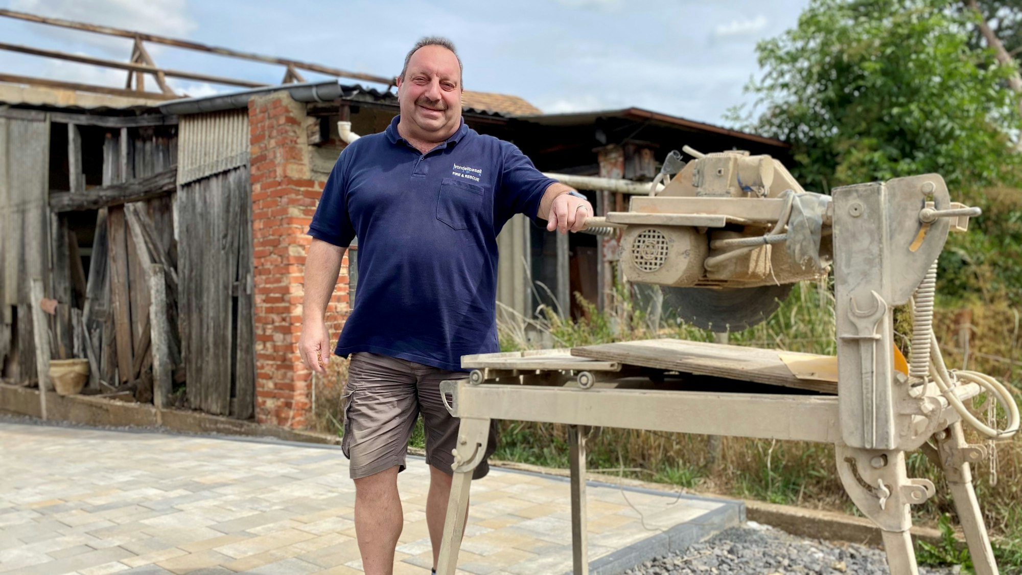 Ein Mann steht in einem Hof an einer Säge, die durch vieles Arbeiten eingestaubt ist.