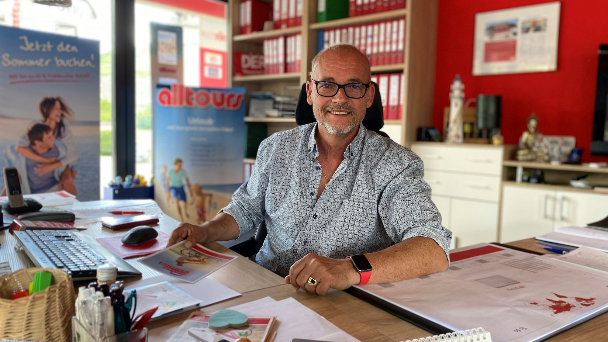 Ein Mann sitzt an einem Schreibtisch in seinem Reisebüro. Im Hintergrund sind Akten und Plakate von Reisen zu sehen.