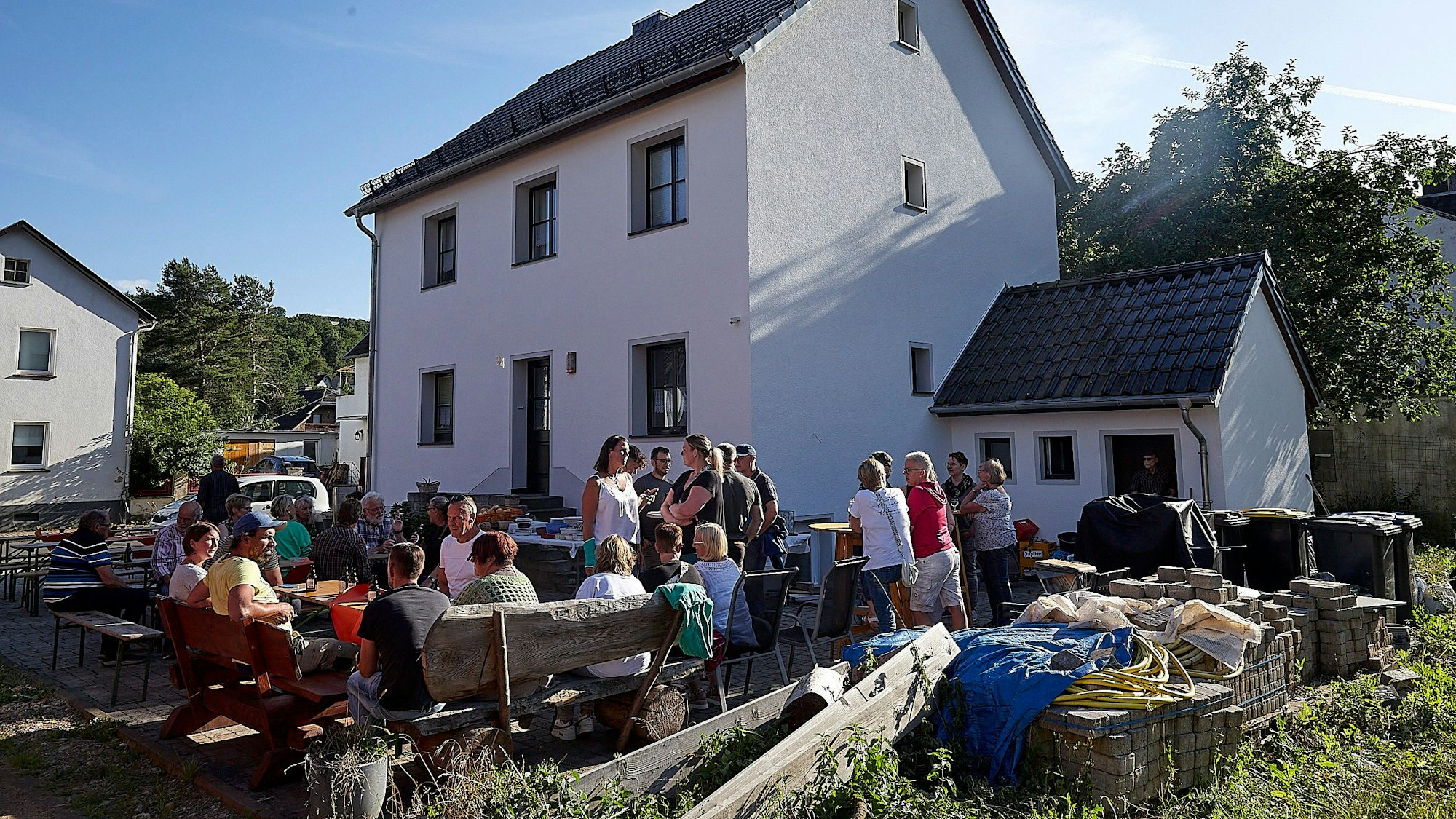 In einem Hof vor einem weißen Haus sitzen zahlreiche Menschen zusammen.