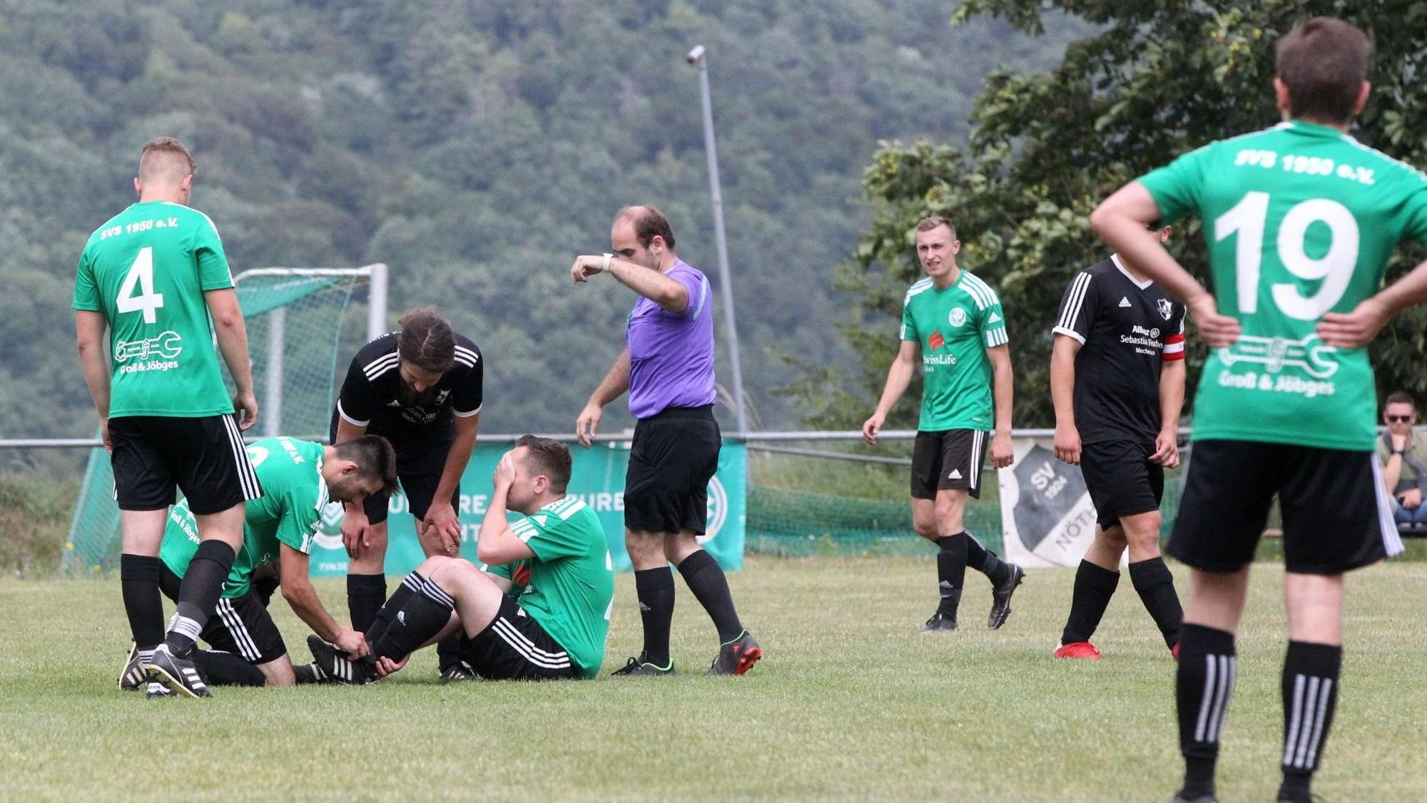 Der Schöneseiffener Spieler Tobias Heinen sitzt auf dem Boden, die Hand vors Gesicht geschlagen. Ein Mitspieler und ein gegnerischer Spieler schauen nach seinem Fuß. Der Schiedsrichter gestikuliert.