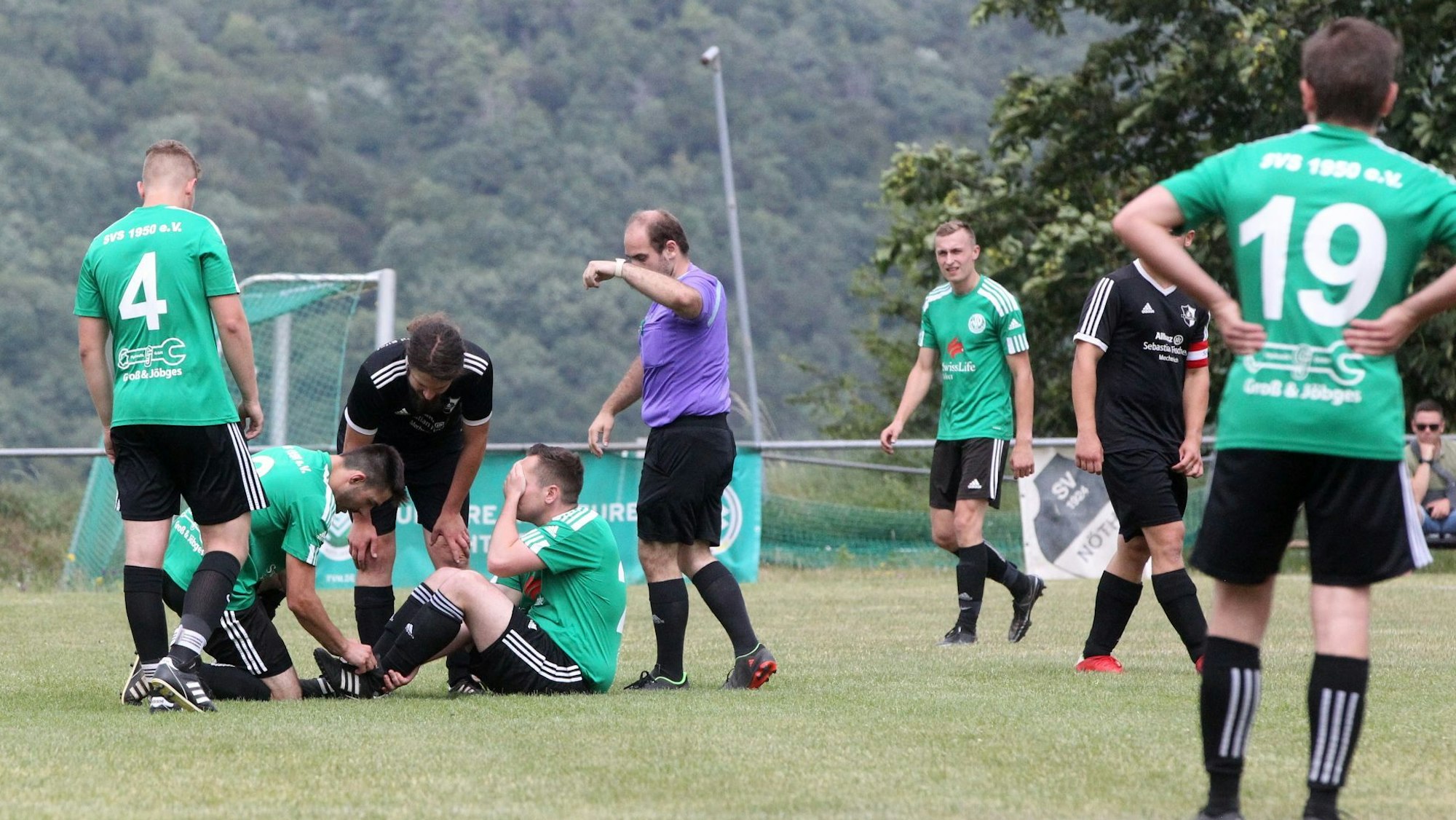 Der Schöneseiffener Spieler Tobias Heinen sitzt auf dem Boden, die Hand vors Gesicht geschlagen. Ein Mitspieler und ein gegnerischer Spieler schauen nach seinem Fuß. Der Schiedsrichter gestikuliert.