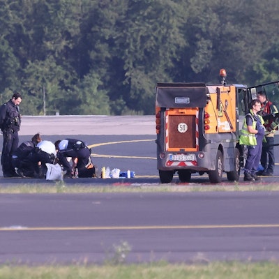 Sicherheitspersonal und Polizisten stehen auf dem Flugfeld und versuchen Aktivisten der Gruppe Letzte Generation vom Asphalt zu lösen, nachdem sie sich festgeklebt haben.