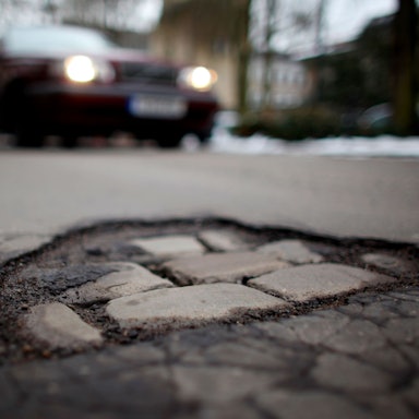 Ein Auto fährt auf ein Schlagloch in der Fahrbahndecke einer Straße in Köln zu.