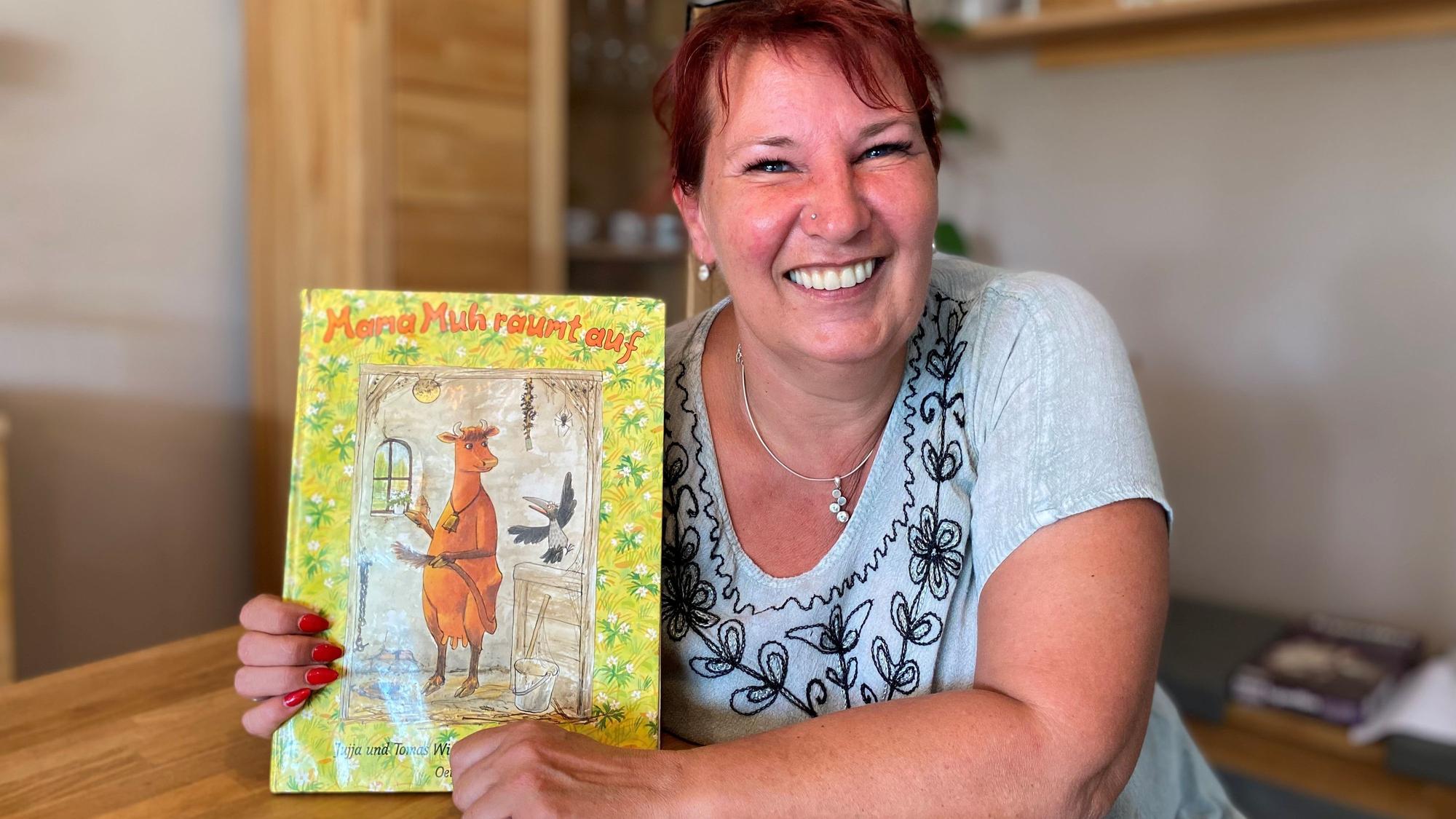 Eine Frau sitzt am Tisch. Sie hält ein Kinderbuch in die Kamera, auf de, eine Kuh abgebildet ist.