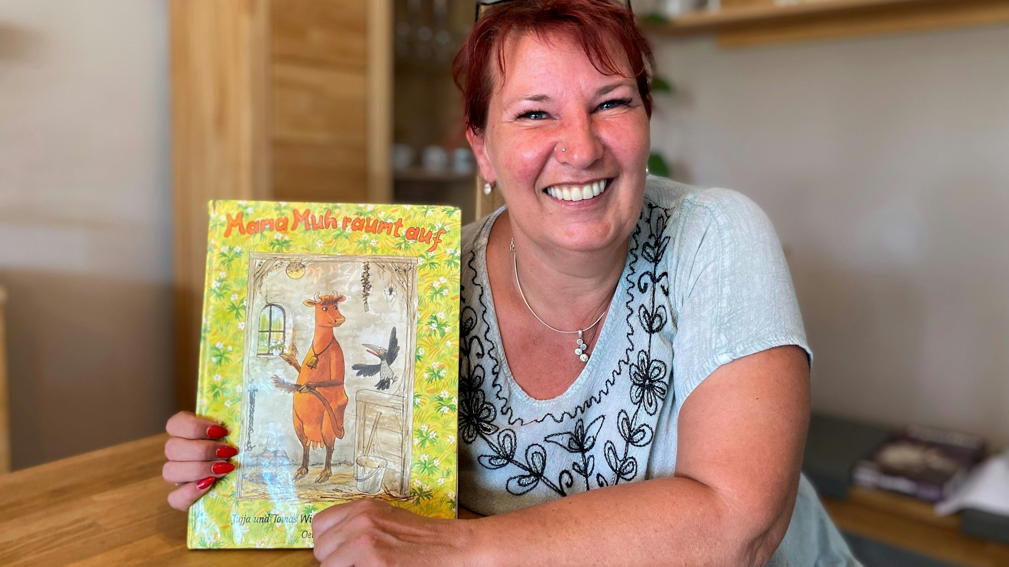 Eine Frau sitzt am Tisch. Sie hält ein Kinderbuch in die Kamera, auf de, eine Kuh abgebildet ist.