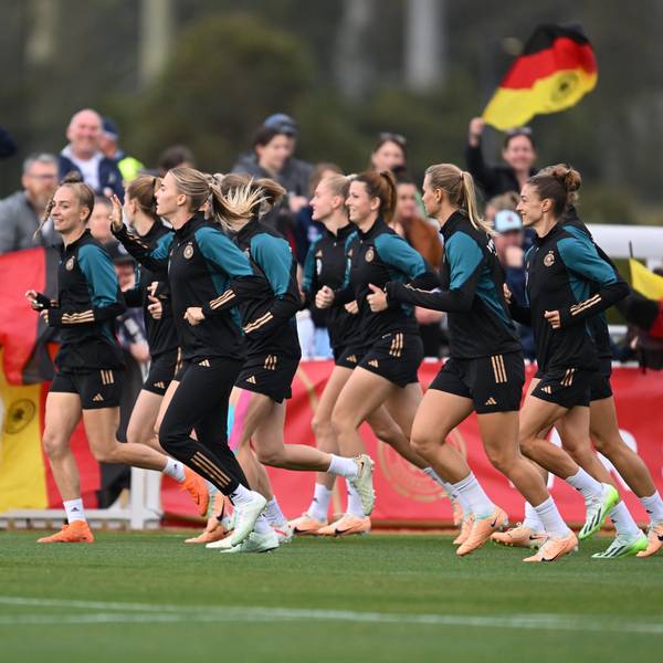 Die deutsche Frauenfußball-Nationalmannschaft wärmt sich während einer offenen Trainingseinheit im Central Coast Regional Sporting & Recreation Centre in Tuggerah auf. Im Hintergrund sind deutsche Fahnen und das Publikum zu sehen.