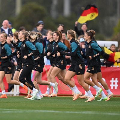 Die deutsche Frauenfußball-Nationalmannschaft wärmt sich während einer offenen Trainingseinheit im Central Coast Regional Sporting & Recreation Centre in Tuggerah auf. Im Hintergrund sind deutsche Fahnen und das Publikum zu sehen.
