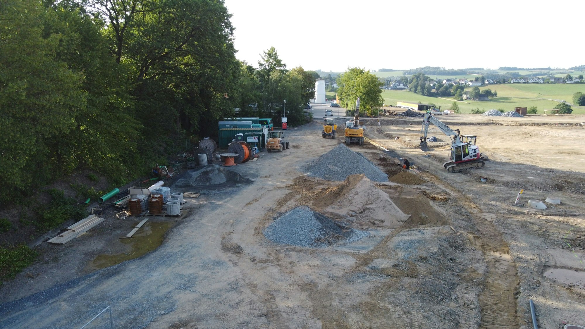 Mehrere Bagger und Baumaterialien auf einer Baustelle. Links Bäume. Im Hintergrund Wiesen und Häuser einer dörflichen Landschaft.
