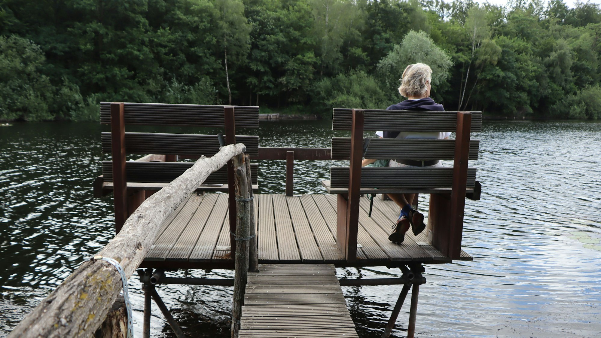 Eine Frau sitzt auf einer Bank, die auf einem Steg steht. Sie lässt den Blick über einen See schweifen.