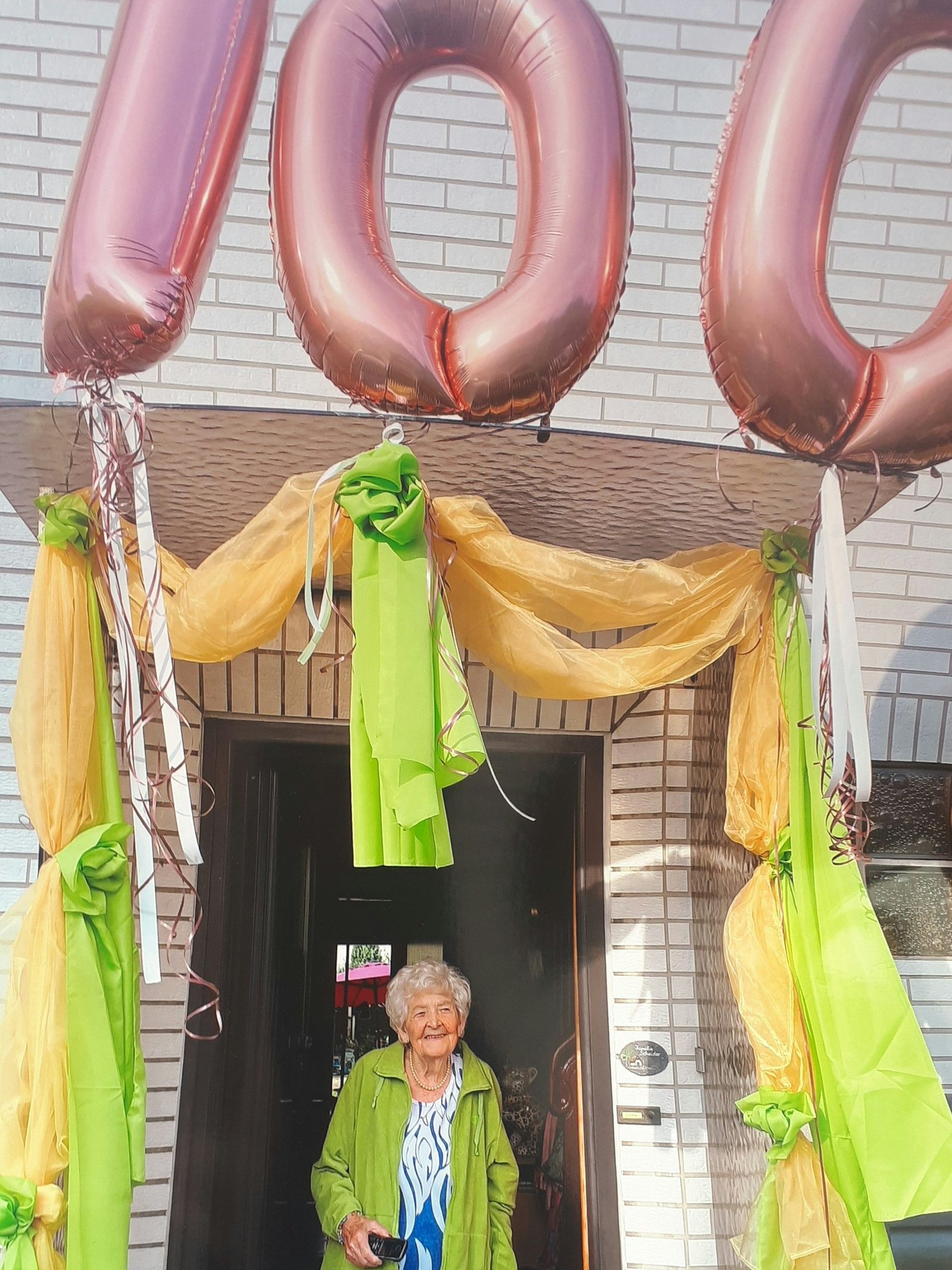 Seniorin, die in einem Türrahmen steht, der mit Luftballons in Form der Zahl „100“ und Tüchern beschmückt ist