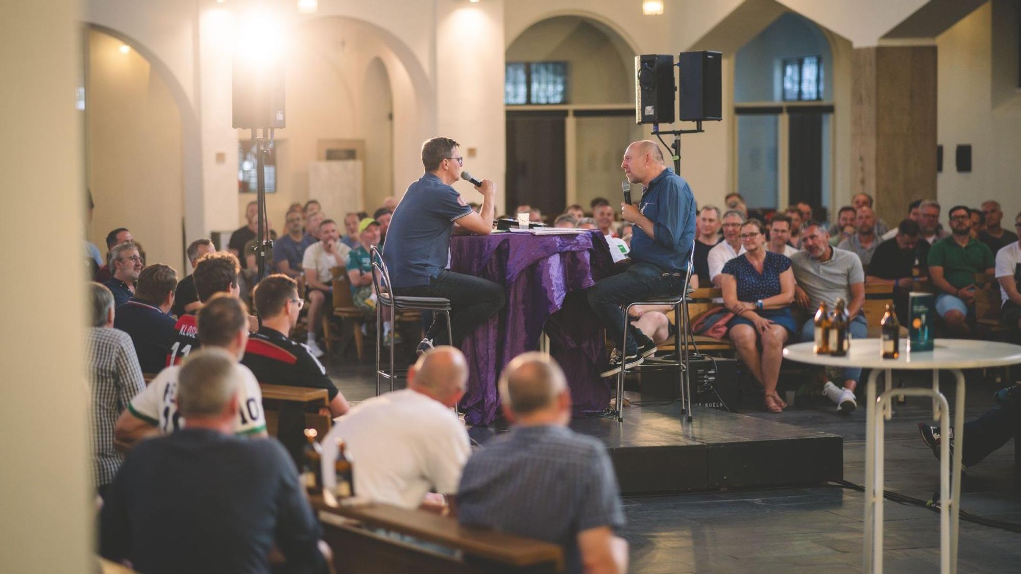 Um ein Podium in der Kirche herum sitzen viele Menschen.