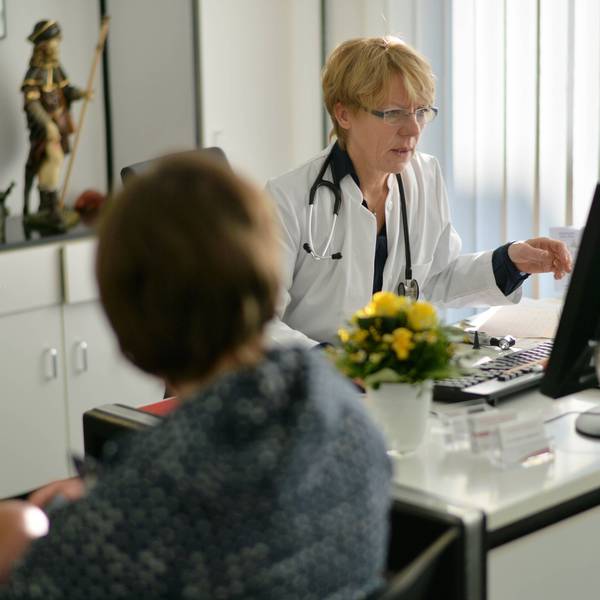 Eine Hausärztin bespricht die Diagnose mit einer Patientin. Sie sitzt am SChreibtisch, trägt einen weißen Kittel und gestikuliert.