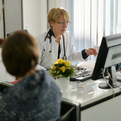 Eine Hausärztin bespricht die Diagnose mit einer Patientin. Sie sitzt am SChreibtisch, trägt einen weißen Kittel und gestikuliert.