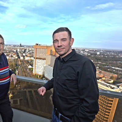 Zwei Männer auf einem Balkon, im Hintergrund sieht man die Skyline von Köln.