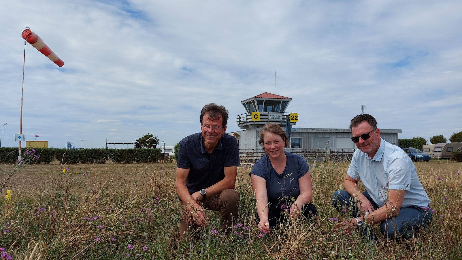 Zwei Männer und eine Frau hocken auf einer Wiese, auf der Blumen blühen. Im Hintergrund sind der Tower eines Flugplatzes und ein Windsack zu sehen.