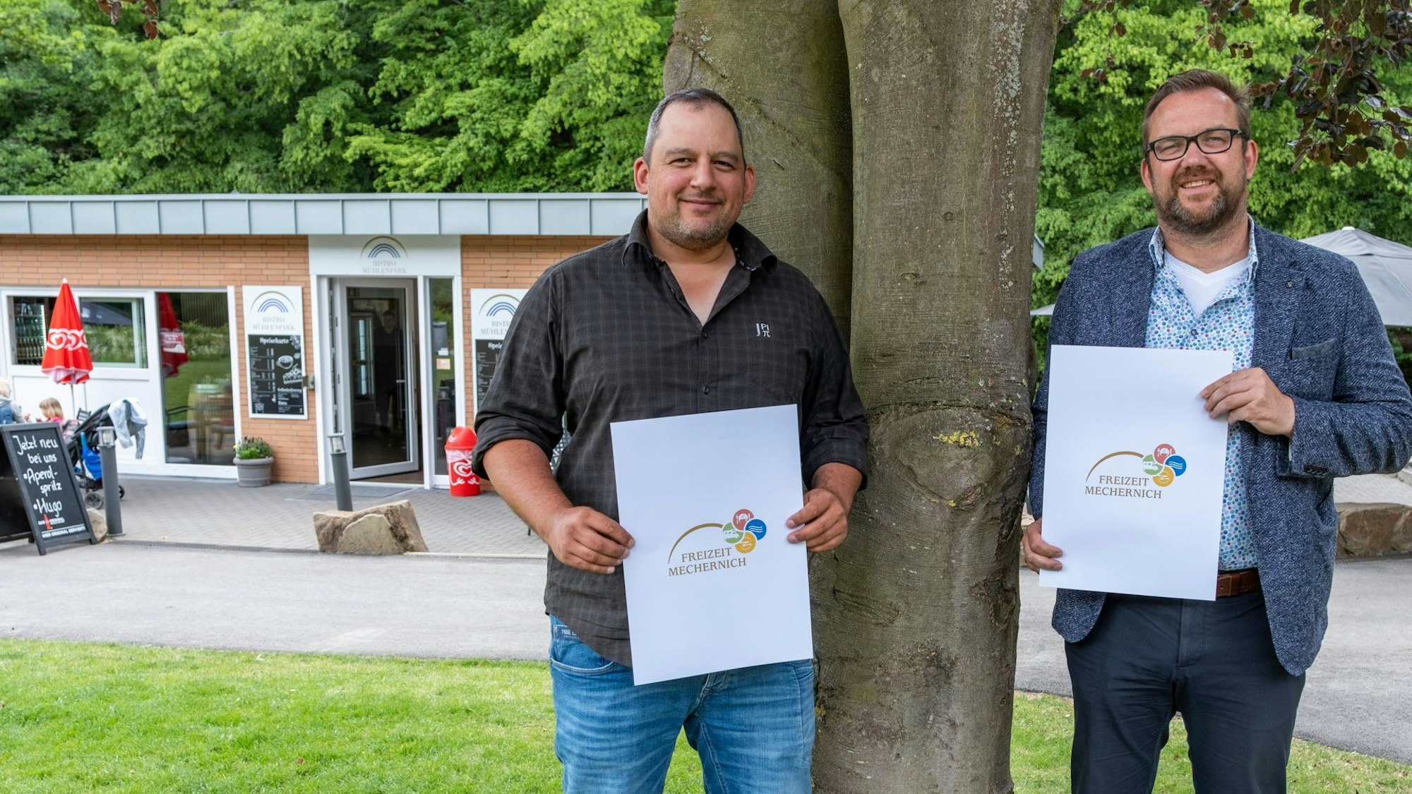 Die beiden Männer stehen an einem Baum und halten Plakate mit einem aufgedruckten Logo in die Kamera.