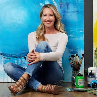 Die Künstlerin Susanne Herbold sitzt vor einer blau-gelben Leinwand, neben ihr stehen Farben und liegen Pinsel.