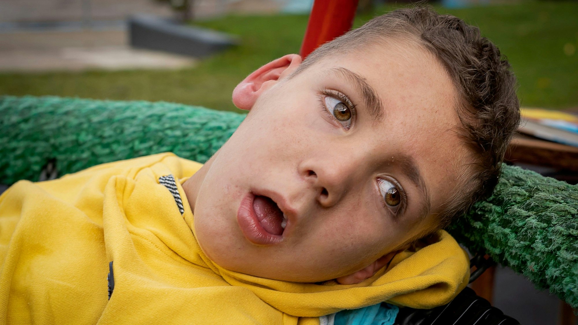 Ein sichtlich an Kinderdemenz erkrankter Junge, bekleidet mit einem gelben Pullover, ist in Großaufnahme zu sehen.