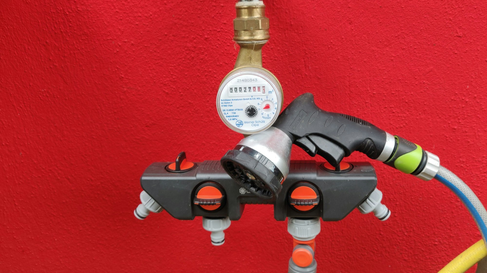 Eine Wasseruhr vor einer roten Hausfassade. An dem Zapfhahn ist ein Gartenschlauch angeschlossen.