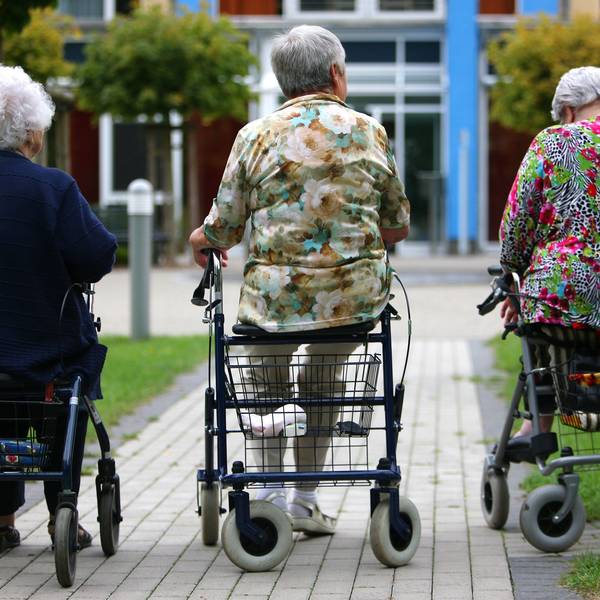 Drei Frauen höheren Alters sitzen auf Rollatoren auf einem Weg, der zu einem Pflegeheim führt