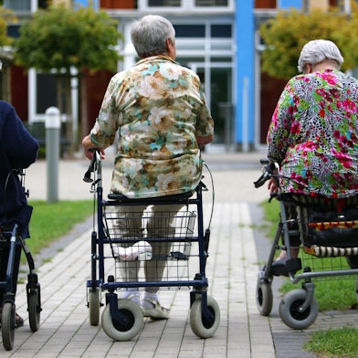 Drei Frauen höheren Alters sitzen auf Rollatoren auf einem Weg, der zu einem Pflegeheim führt