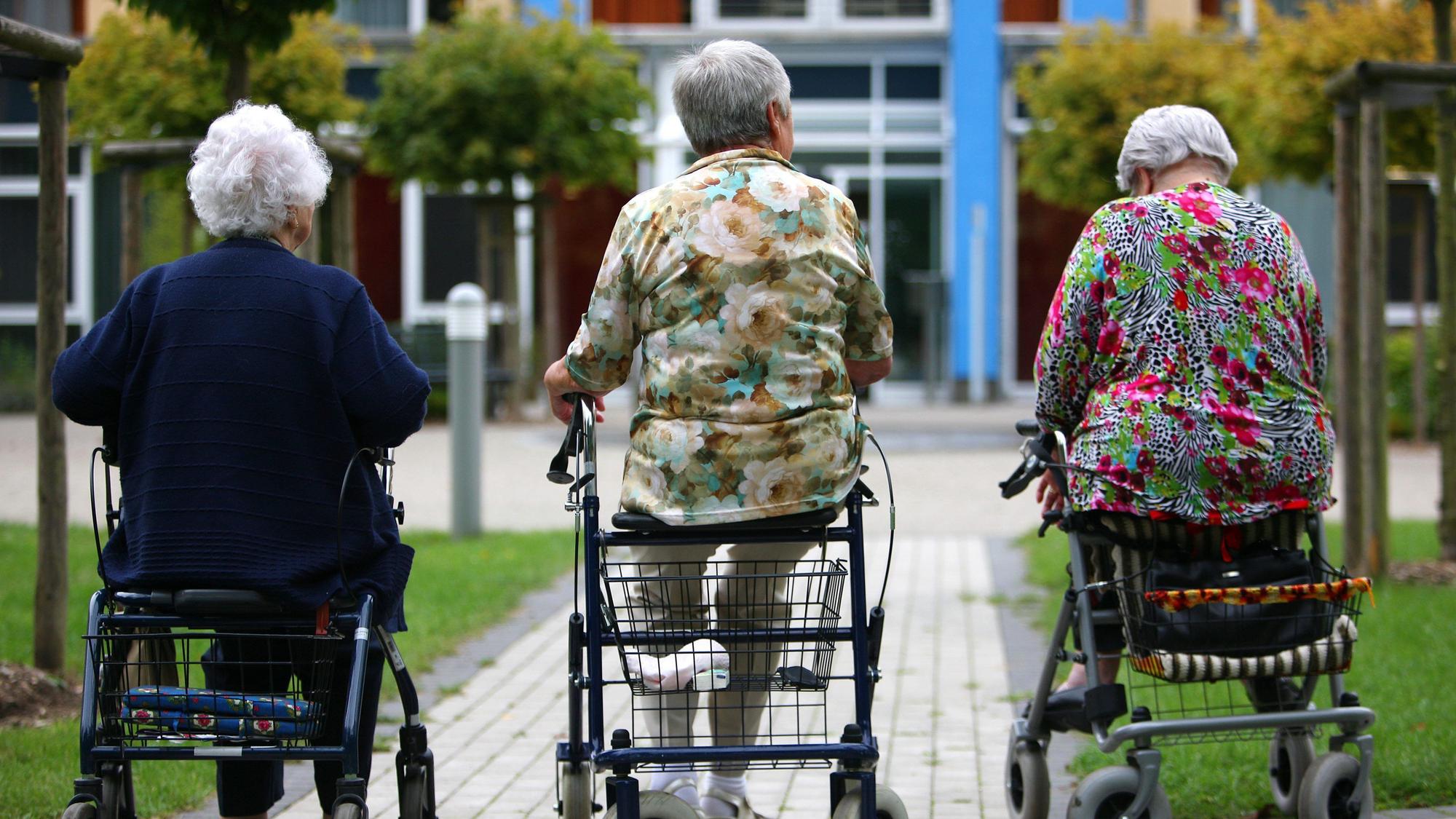 Drei Frauen höheren Alters sitzen auf Rollatoren auf einem Weg, der zu einem Pflegeheim führt