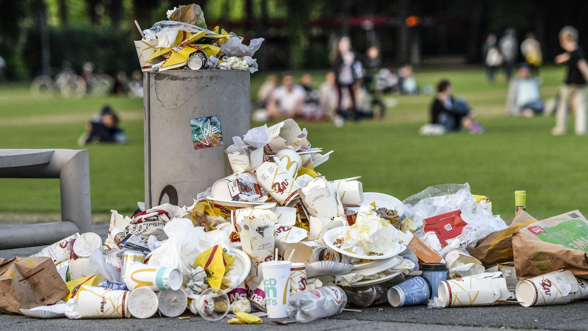 Vor einem bis oben gefüllten Mülleimer liegen Berge von Plastikbechern und anderer Verpackungsmüll. Im Hintergrund befinden sich Menschen auf einer grünen Wiese.