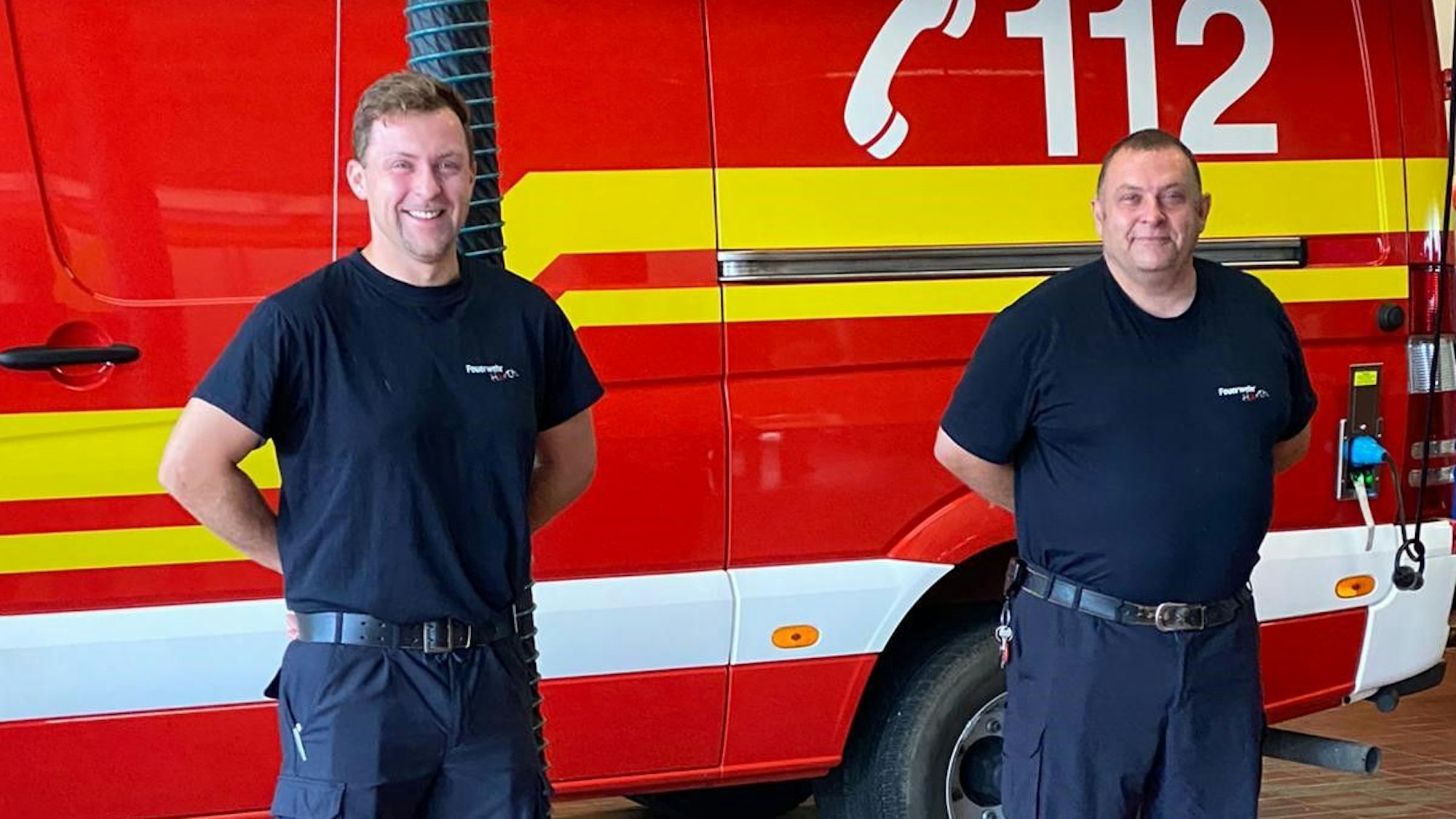 Zwei Männer in dunkelblauer Feuerwehr-Kleidung stehen, die Arme hinter dem Rücken verschränkt, vor einem roten Einsatzwagen.