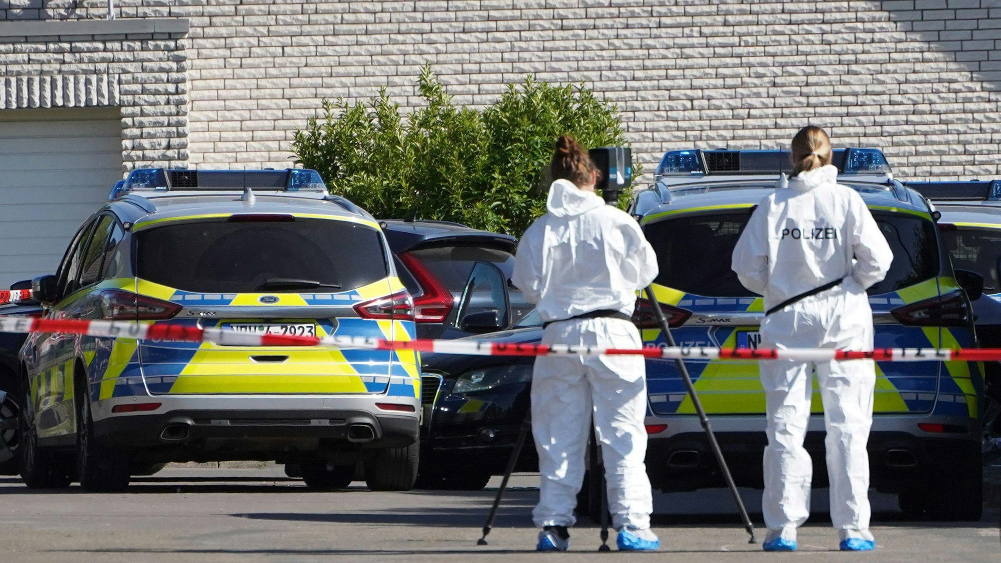 Beamte der Spurensicherung ermitteln an einem Tatort. Sie tragen weiße Anzüge, im Hintergrund sind mehrere Polizeifahrzeuge zu sehen.