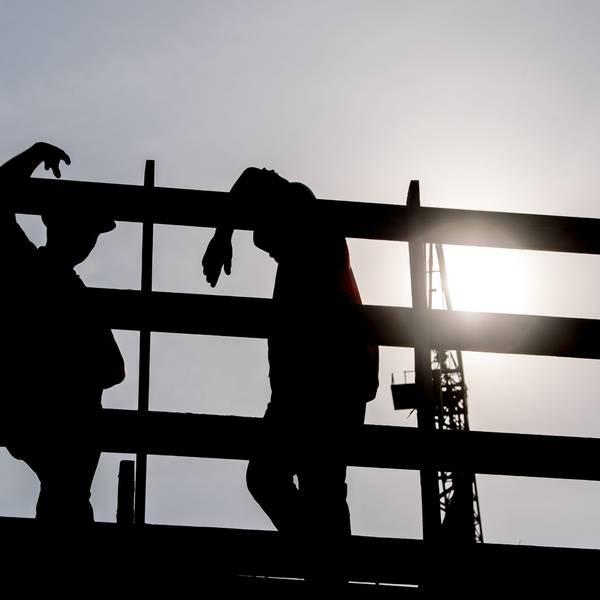 Im Gegenlicht der Morgensonne stehen zwei Arbeiter auf einem Gerüst auf einer Baustelle und unterhalten sich.