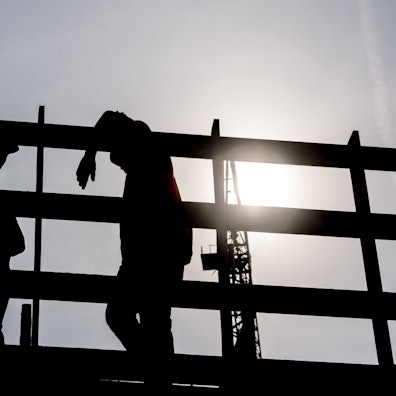 Im Gegenlicht der Morgensonne stehen zwei Arbeiter auf einem Gerüst auf einer Baustelle und unterhalten sich.