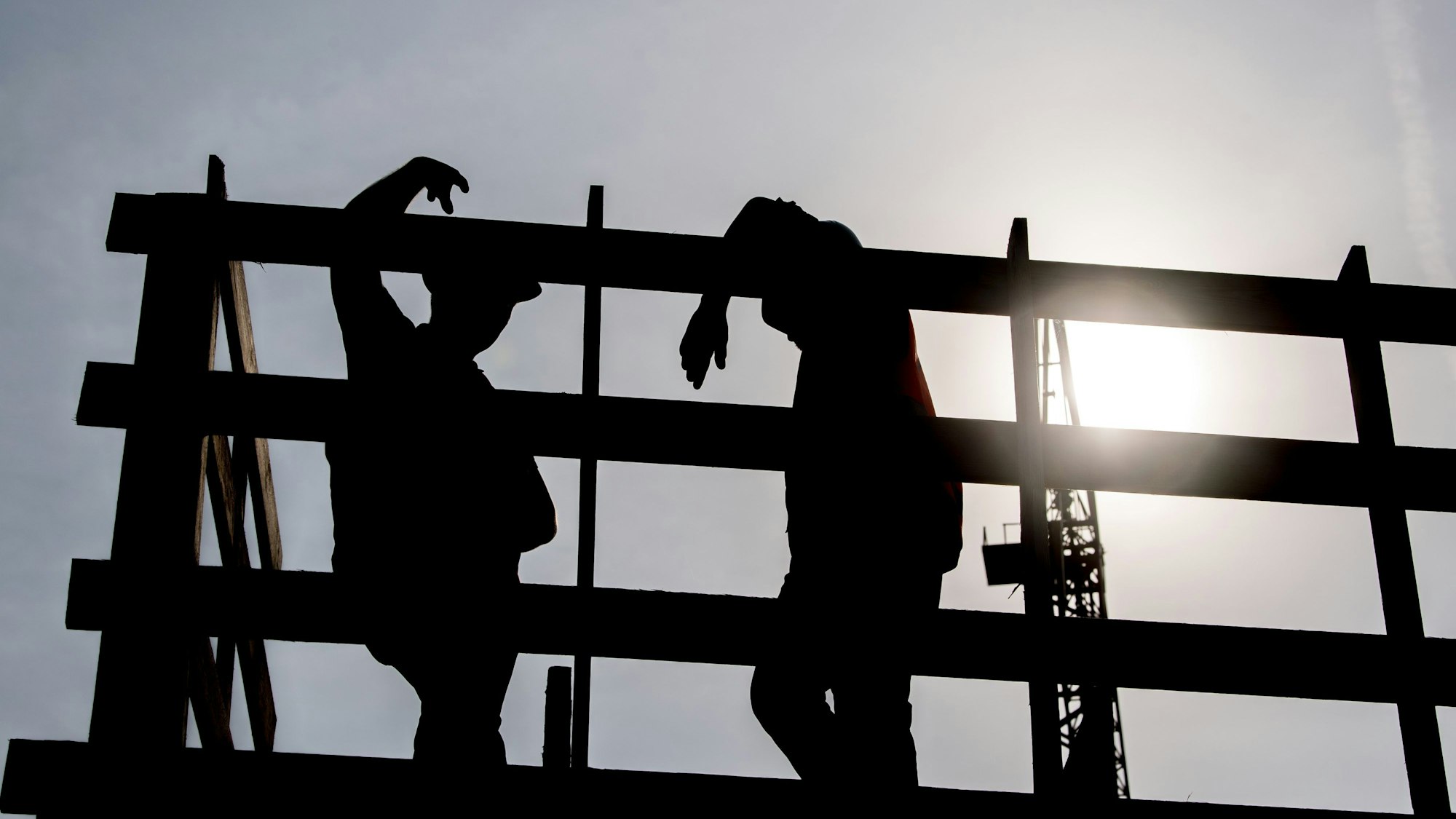 Im Gegenlicht der Morgensonne stehen zwei Arbeiter auf einem Gerüst auf einer Baustelle und unterhalten sich.