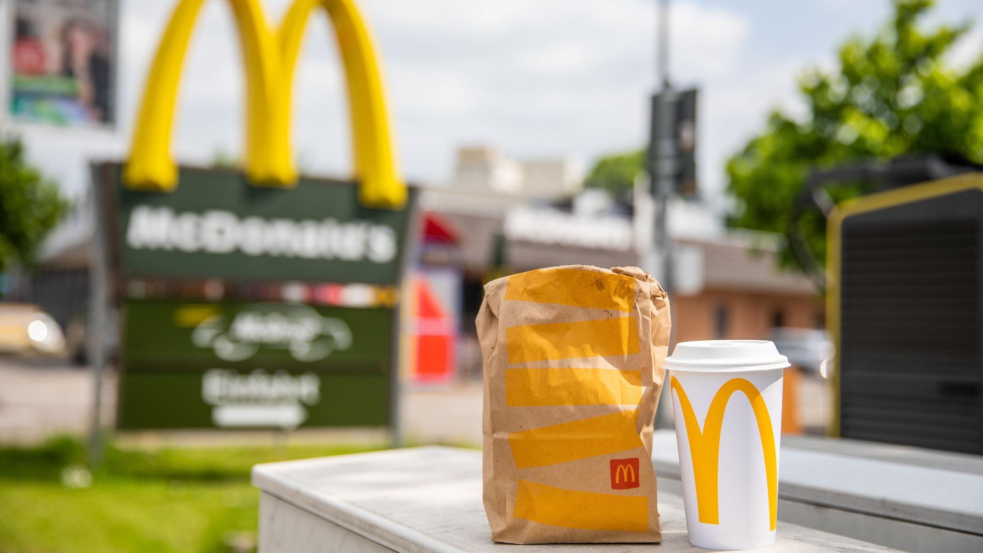 Eine McDonald's To-Go-Tüte und ein dazugehöriger To-Go-Becher stehen vor McDonald's Filiale.