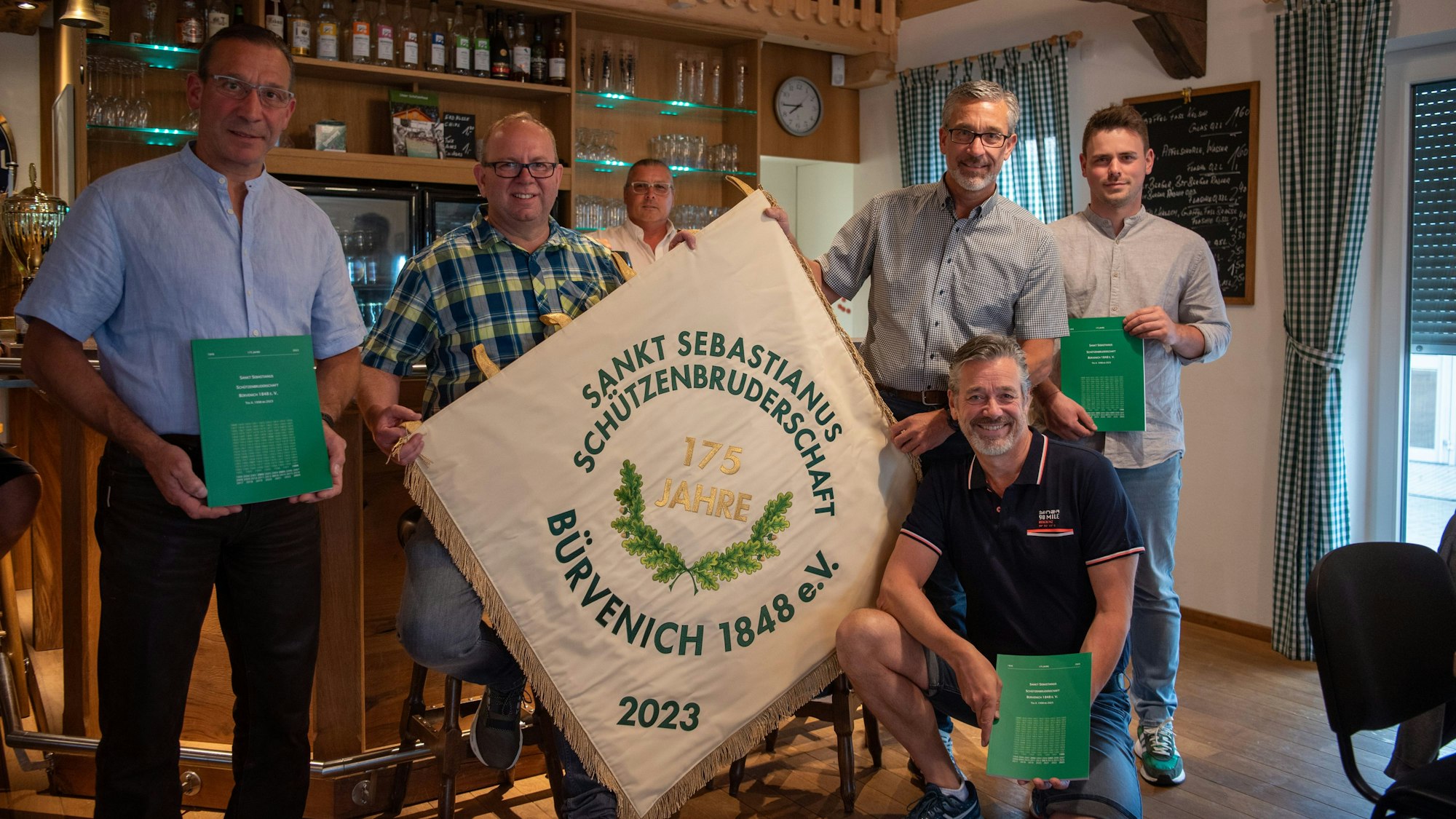 Sechs Männer stehen vor einer Theke und halten eine Fahne hoch.