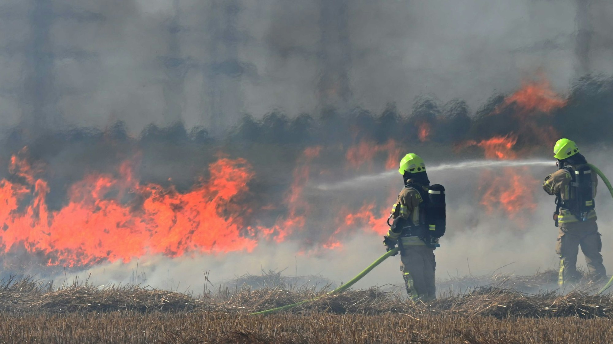 Das Bild zeigt zwei Feuerwehrmänner, die mit Wasser gegen die Flammen ankämpfen.