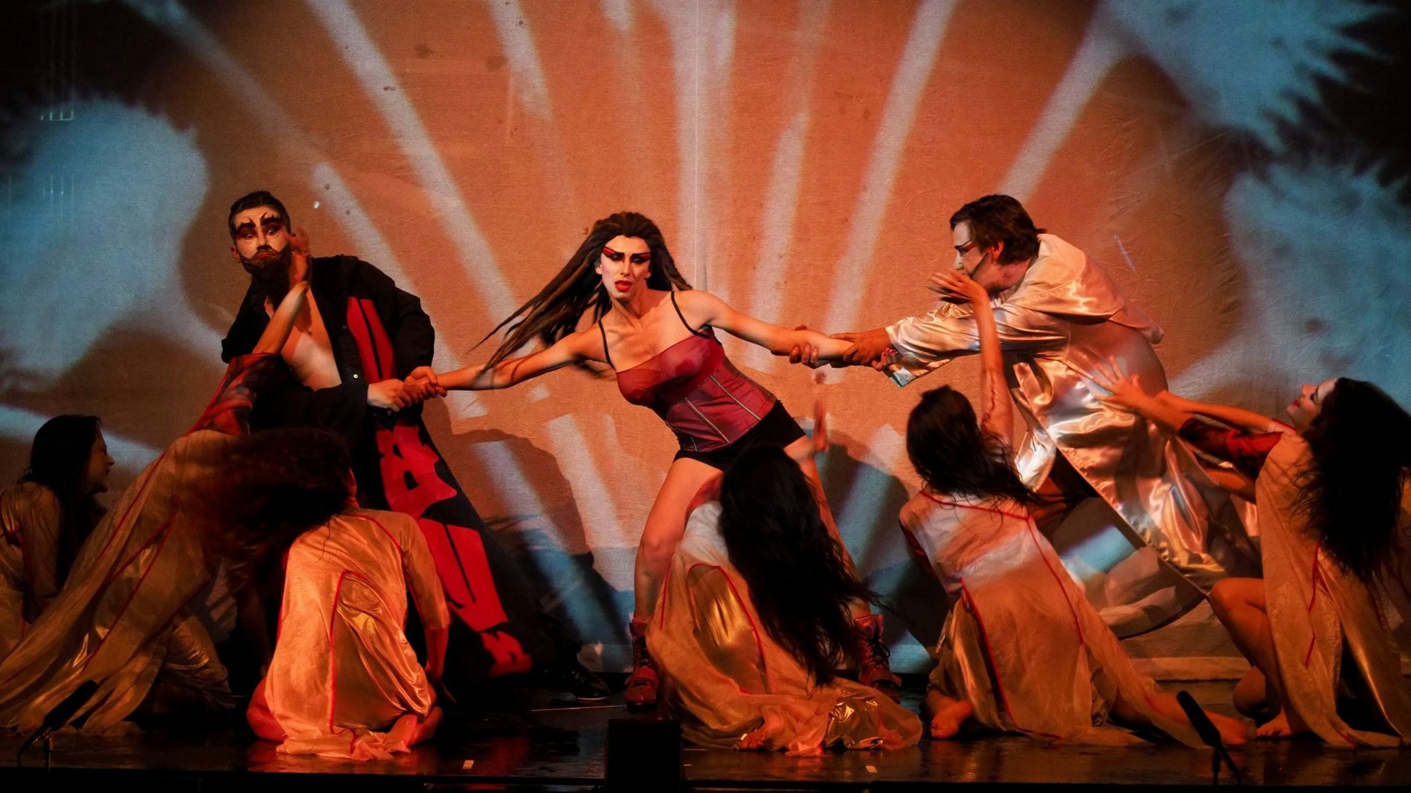 Zwei Herren zerren an der Protagonistin herum, sie trägt ein Korsett und hat Dreadlocks, ein weiß geschminktes Gesicht. Zu ihren Füßen viele flehende Damen.