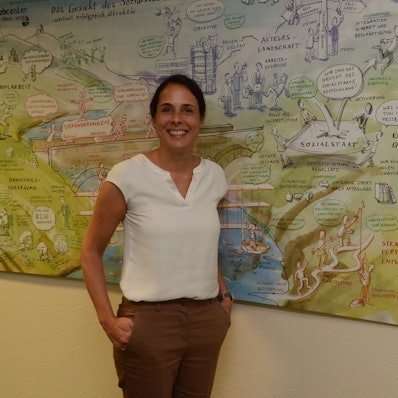 Eine Frau mit dunklen Haaren, weißer Bluse und brauner Hose steht vor einem Bild, das den Sozialstaat erklärt. Sie lächelt den Fotografen an.