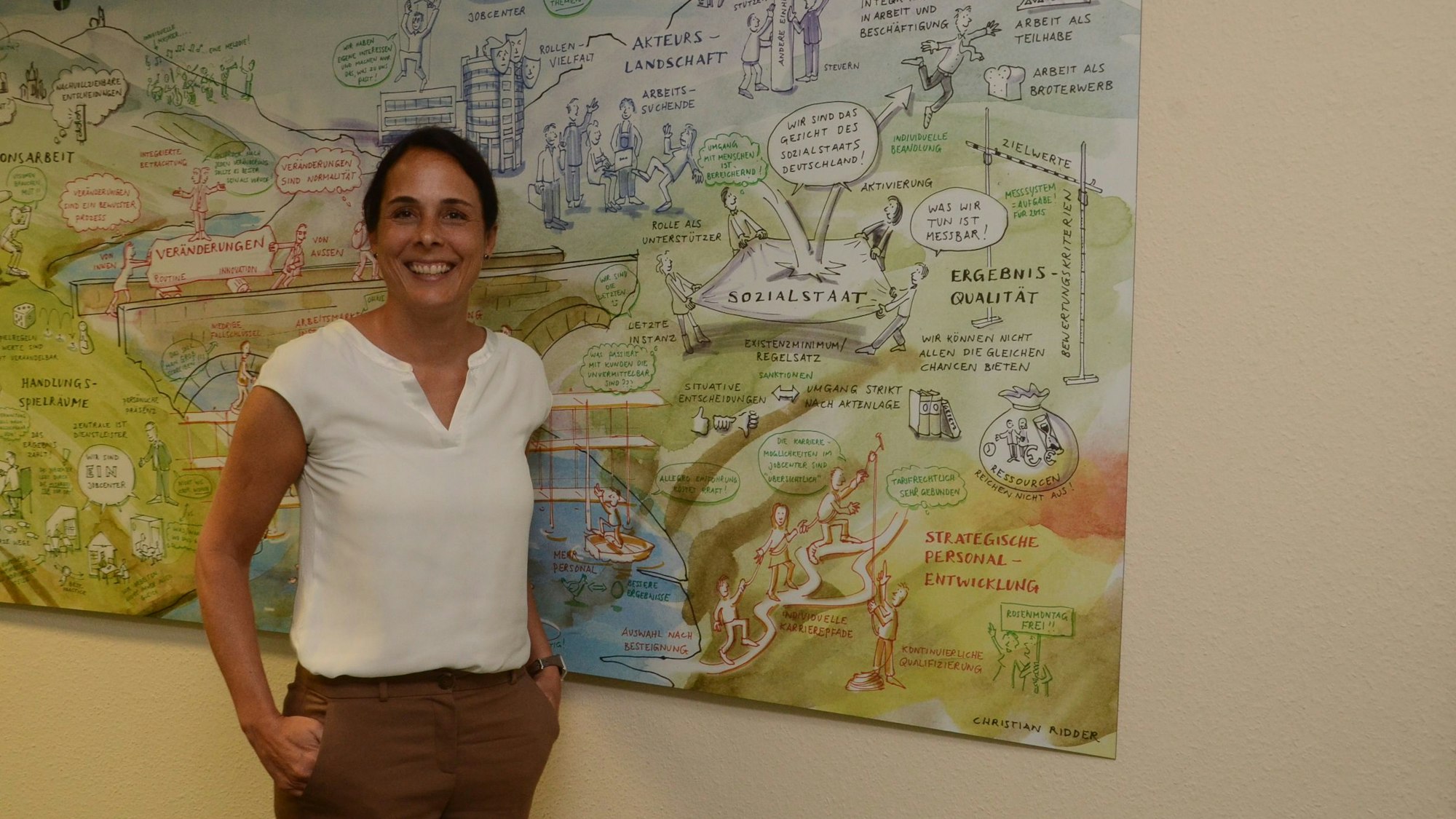 Eine Frau mit dunklen Haaren, weißer Bluse und brauner Hose steht vor einem Bild, das den Sozialstaat erklärt. Sie lächelt den Fotografen an.