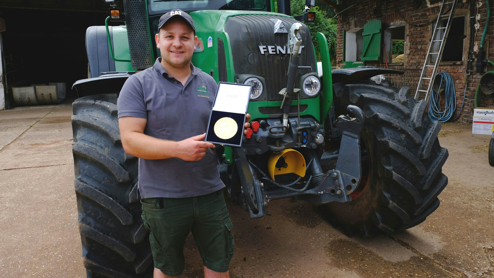 Ein junger Mann steht vor einem Traktor und präsentiert eine goldene Medaille.