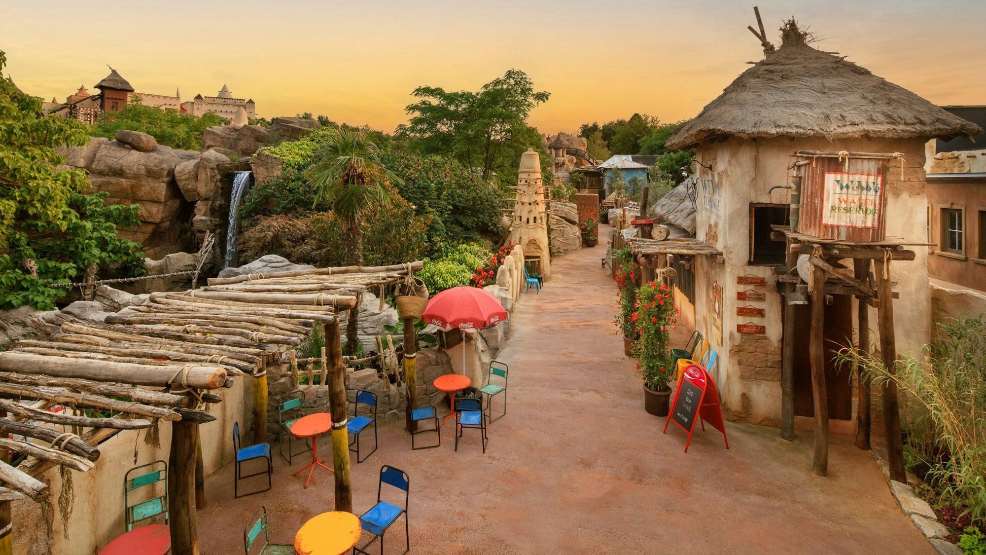 Ein Weg führt zwischen sandfarbenen Häusern hindurch. Links stehen bunte Tische und Stühle. Im Hintergrund ist das Hotel Matamba des Phantasialands zu sehen.
