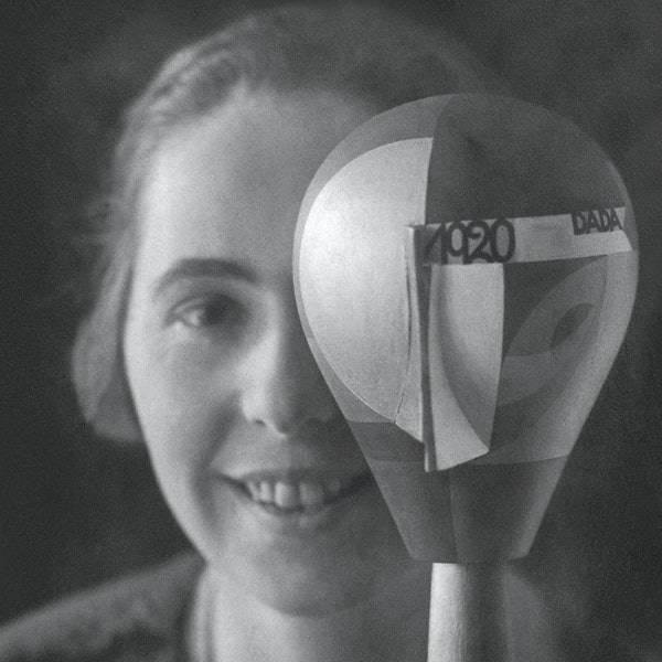 Sophie Taeuber-Arp schaut in dei Kamera, ein Auge wird von einem beklebten Puppenkopf verdeckt.