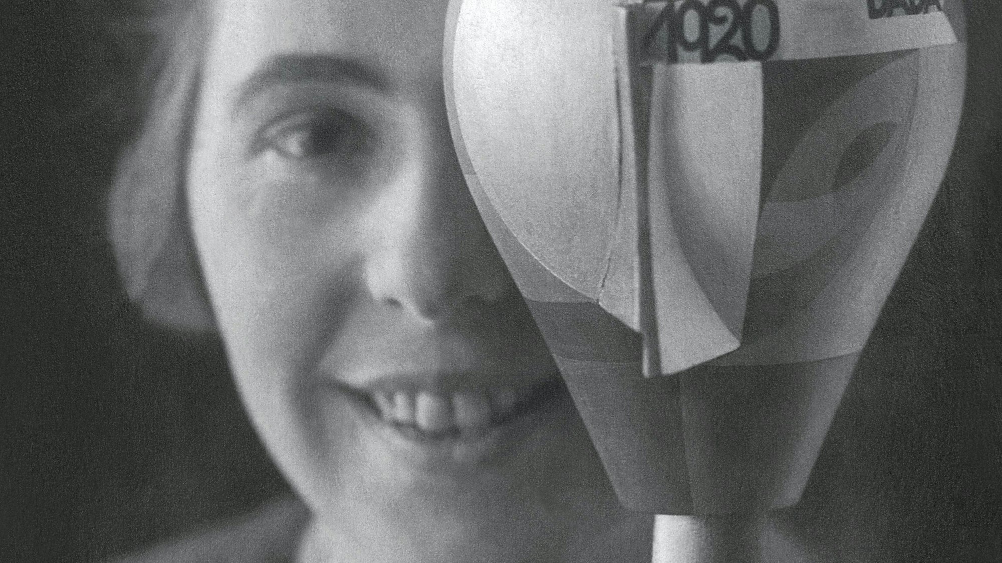 Sophie Taeuber-Arp schaut in dei Kamera, ein Auge wird von einem beklebten Puppenkopf verdeckt.