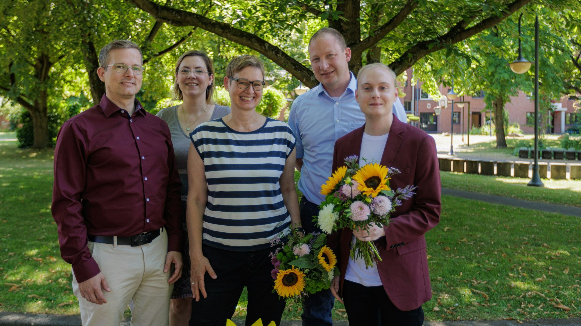 Drei Männer und zwei Frauen stehen im Grünen, zwei halten Blumensträuße.