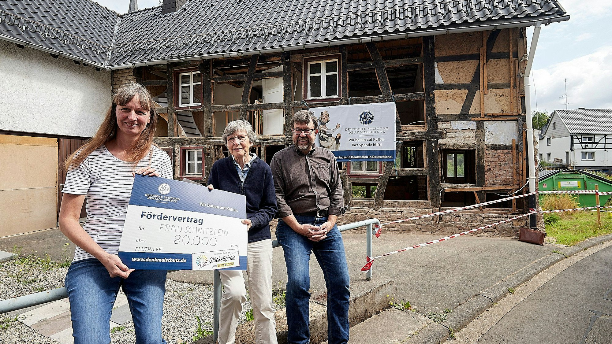 Zwei Frauen und ein Mann sitzen auf einem Geländer vor einem verfallenen Haus. Die Frauen halten einen großen Scheck über 80.000 Euro in der Hand.