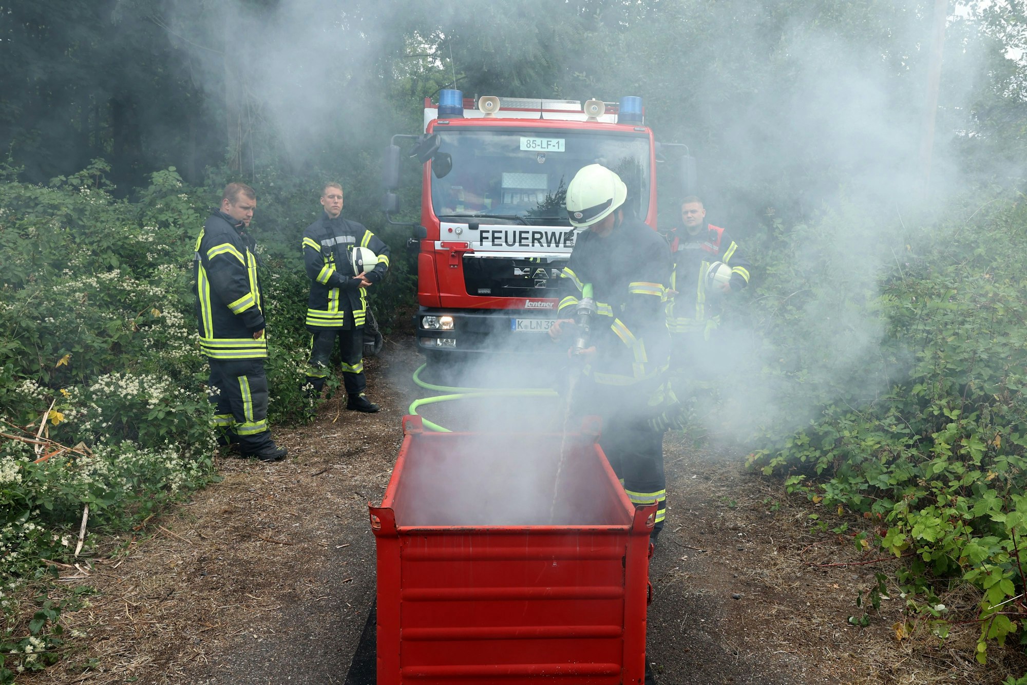 Mehrere Einsatzkräfte der Feuerwehr stehen im Wald. Ein Feuerwehrmann löscht einen Brand in einer roten Box.