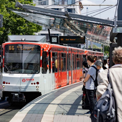 Eine rot-weiße Straßenbahn an einer Haltestelle. Menschen warten auf den Zug.