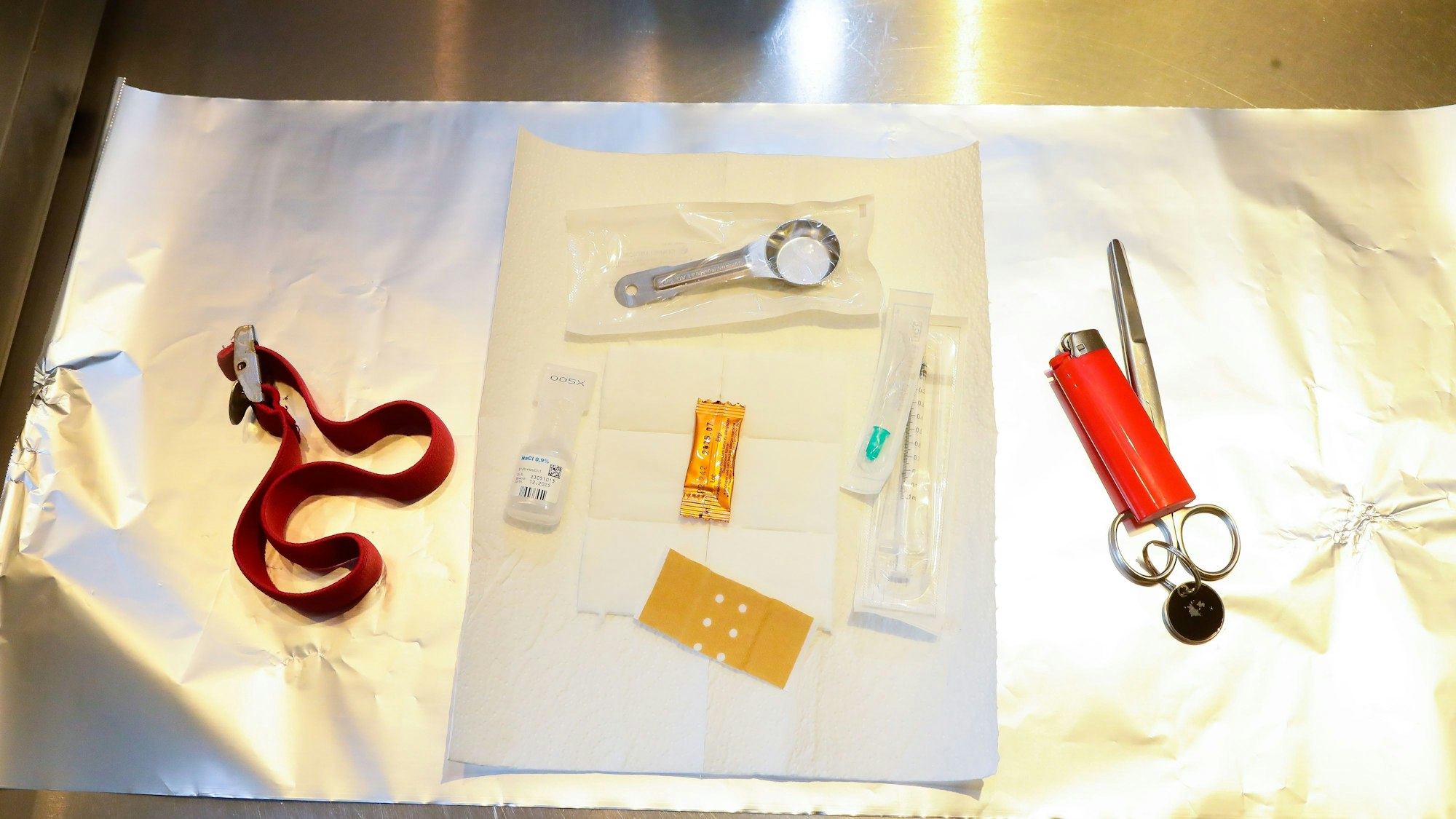 Auf einer Alufolie liegen ein Venenstauer, eine Schere, ein Feuerzeug, ein Pflaster, eine Spritze, ein Löffel, Vitamin C, Kochsalzlösung und ein Filter zum Konsumieren von Heroin.