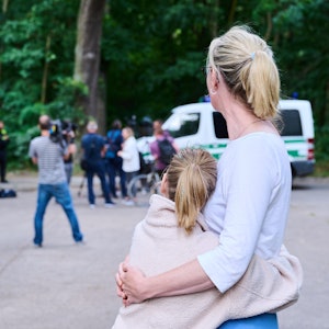 Eine Anwohnerin und ihre Tochter sehen dem Treiben von Journalisten und Polizei zu, während Tierexperten und Polizeibeamten in einem Waldgebiet in Zehlendorf stehen. Eine frei laufende Raubkatze sollte in der Gemeinde Kleinmachnow in Brandenburg gesichtet worden sein. Am Freitag ist die Suche eingestellt worden.